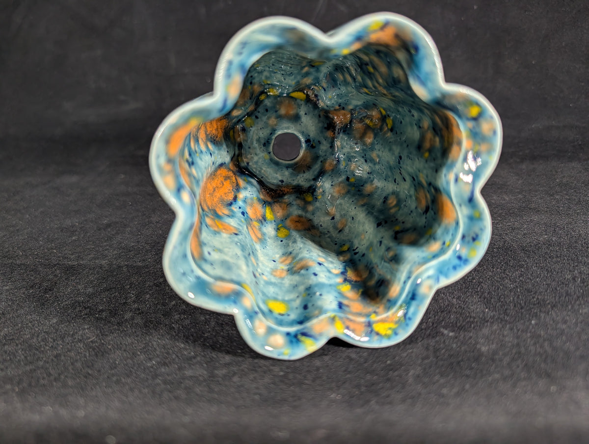 A handmade ceramic flower-shaped bowl with a scalloped rim, featuring a marbled glaze of blue, orange, yellow, and dark brown on a dark gray background.