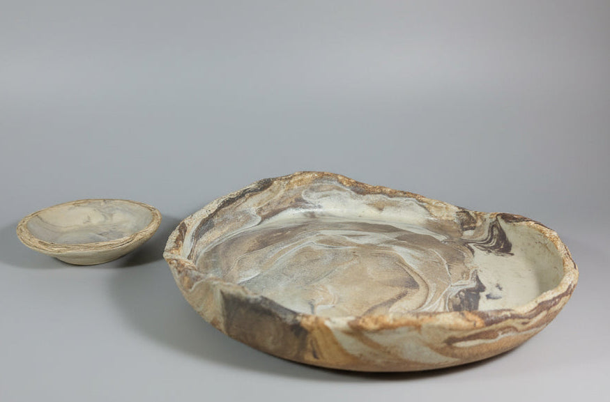 Two hand-carved stone plate and dipping dish with natural marbled patterns in beige, brown, and gray, displayed on a neutral gray studio background.