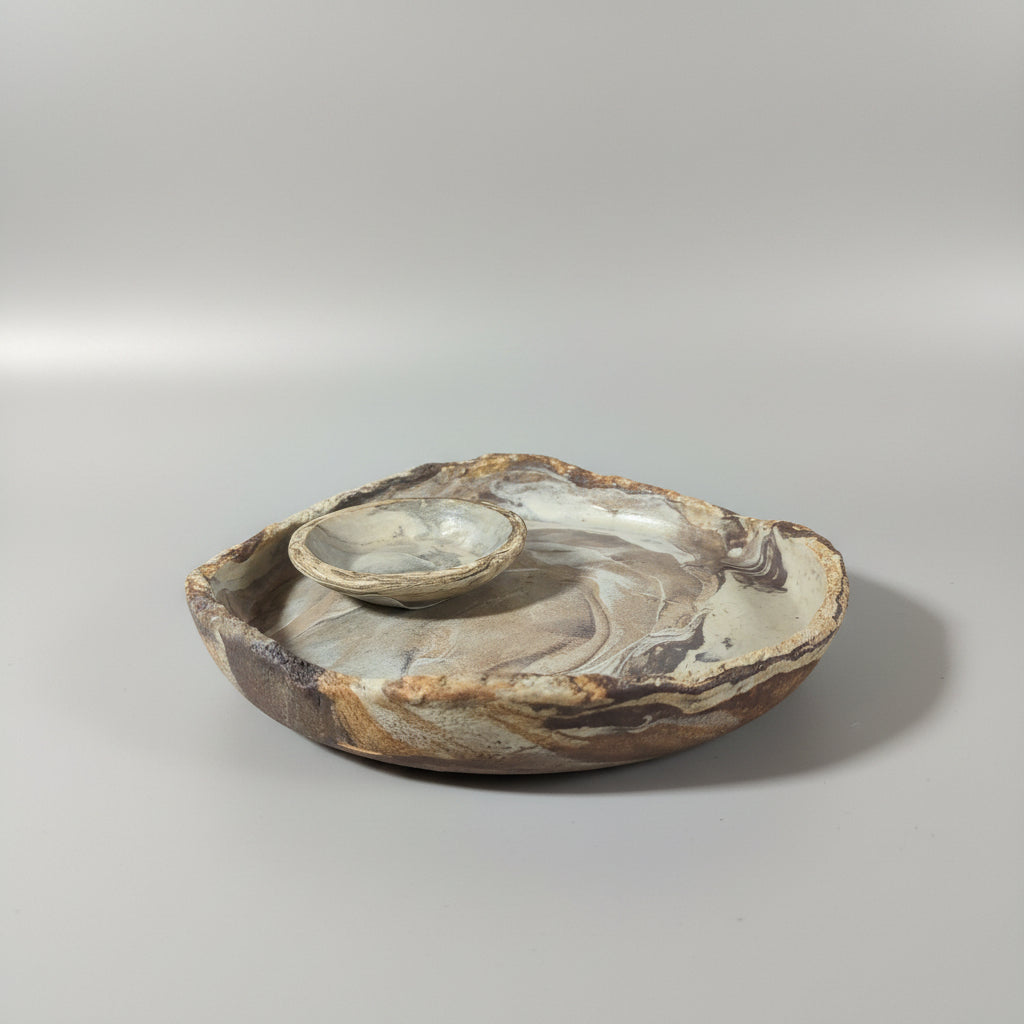 Two handmade ceramic plate and dipping dish with a swirled marbled pattern in beige, brown, and gray, featuring a small raised circular element on top, set against a dark background.